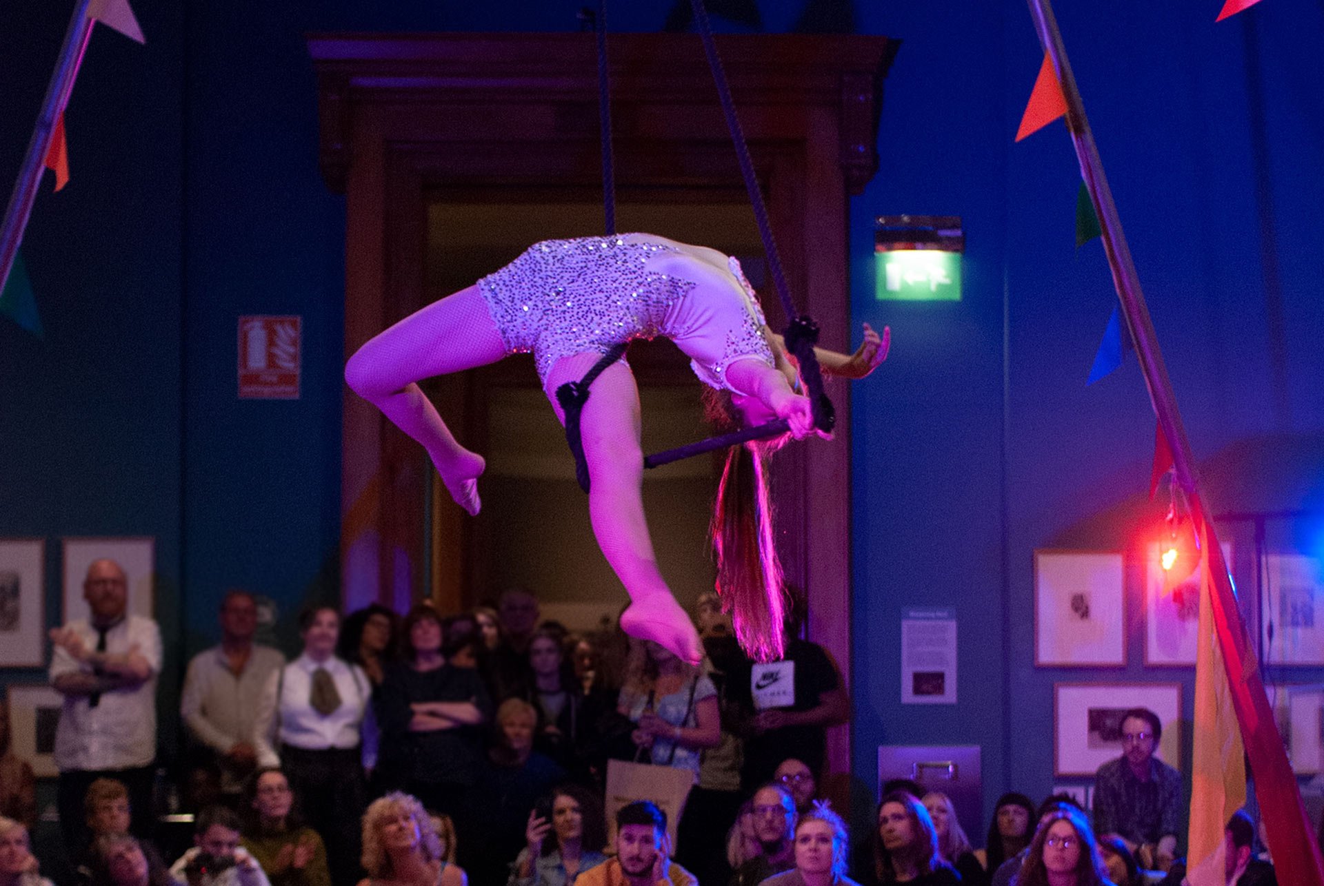 a-circus-gymnast-spinning-in-the-air-while-hanging-on-a-rope-with-a-rim-in-the-Weston-Park-Gallery-in-Sheffield-UK-with-an-audience-surrounding-her