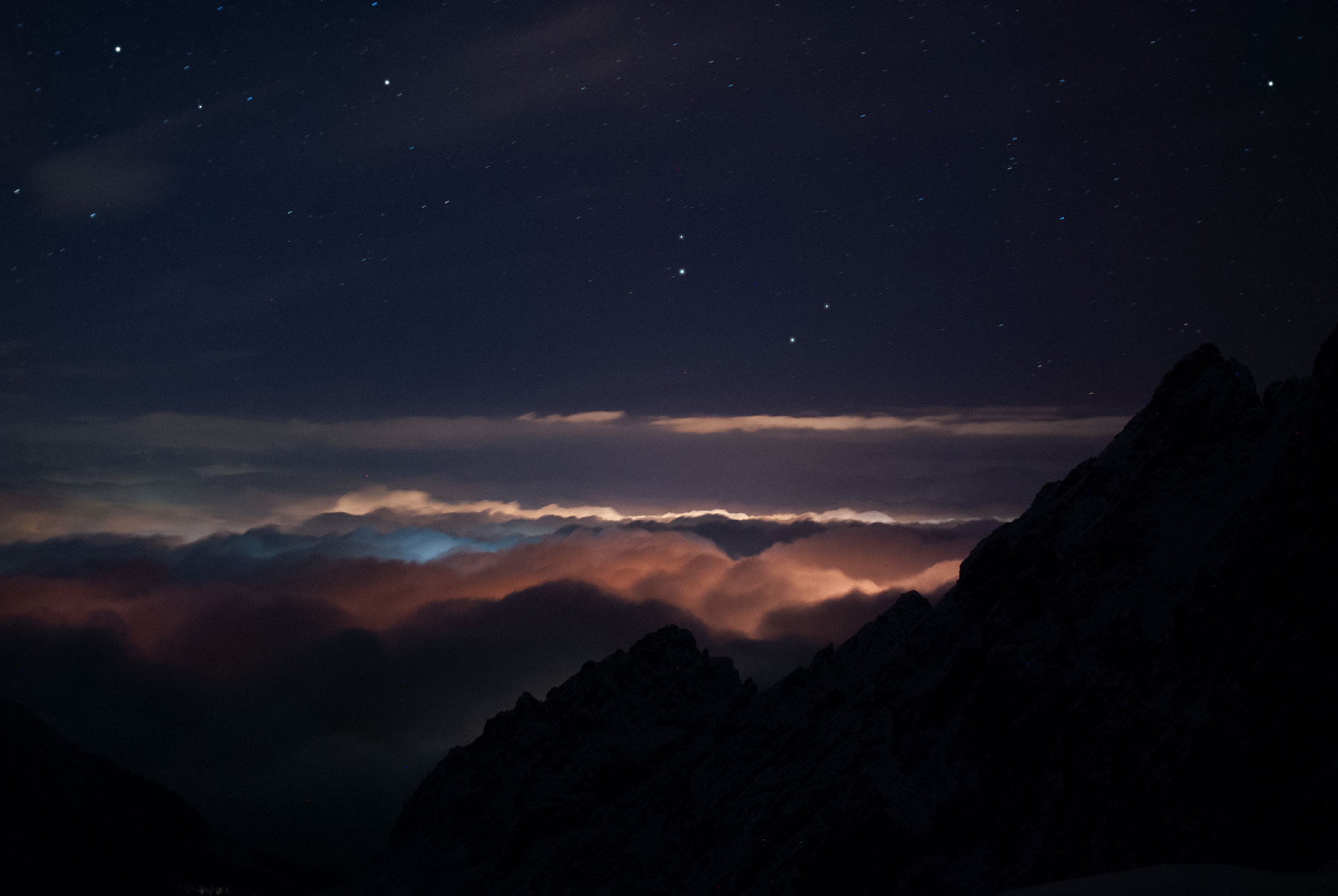 a-photograph-from-heigh-dark-mountains-of-a-small-town-that-rests-under-the-clouds-illuminating-them